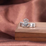 Bague claddagh en argent avec un motif de mains tenant une coeur qui porte une couronne posée sur une planche en bois.