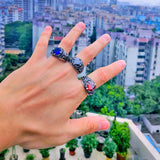 Main d'homme portant une bague bleue, une bague noire et une bague rouge, avec un arrière-plan urbain.