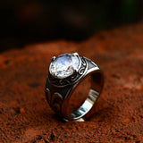 Bague pour homme posée sur une surface en terre, avec une pierre blanche.