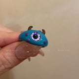 Bague créature bleue avec un seul œil bleu, des tâches violettes et deux cornes noires. 