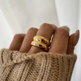 Main de femme portant une bague dorée en acier inoxydable, au design épais formant deux anneaux collés.