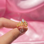 Bague plaqué or en forme de couronne, ornée de zircons colorés, tenue délicatement entre deux doigts.